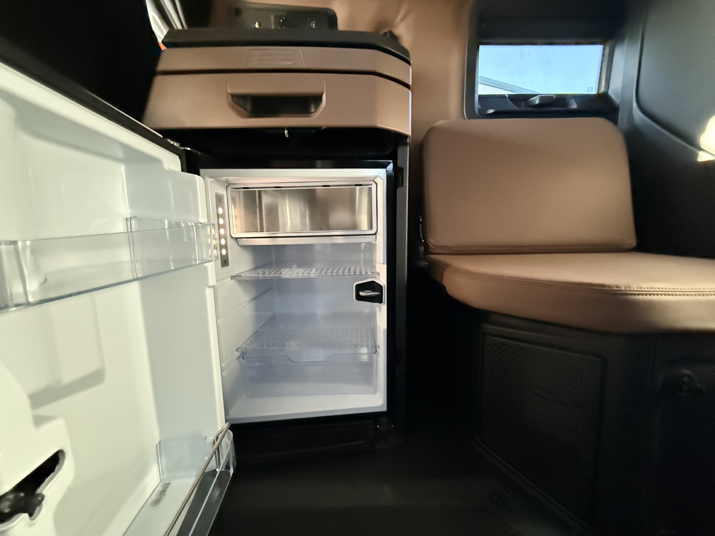Cab interior of a 2026 Mack PR64T76H showing an open mini-fridge with glass door and empty shelves, small storage compartment above, leather seat and dark interior trim.
