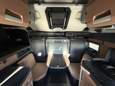 Mack PR64T76H cab interior showing sleeper berth with tan upholstery, center console, overhead storage, and cockpit area.