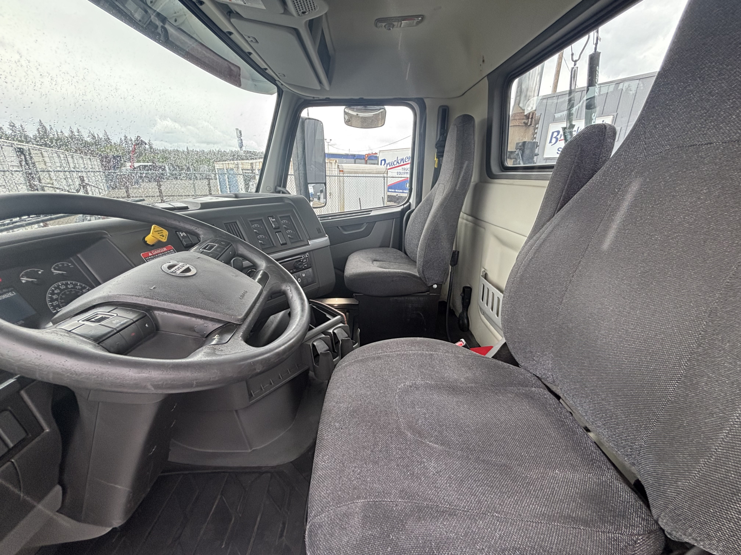 Truck cab interior showing steering wheel and dashboard of a Volvo VNL84T300 with grey fabric seats.