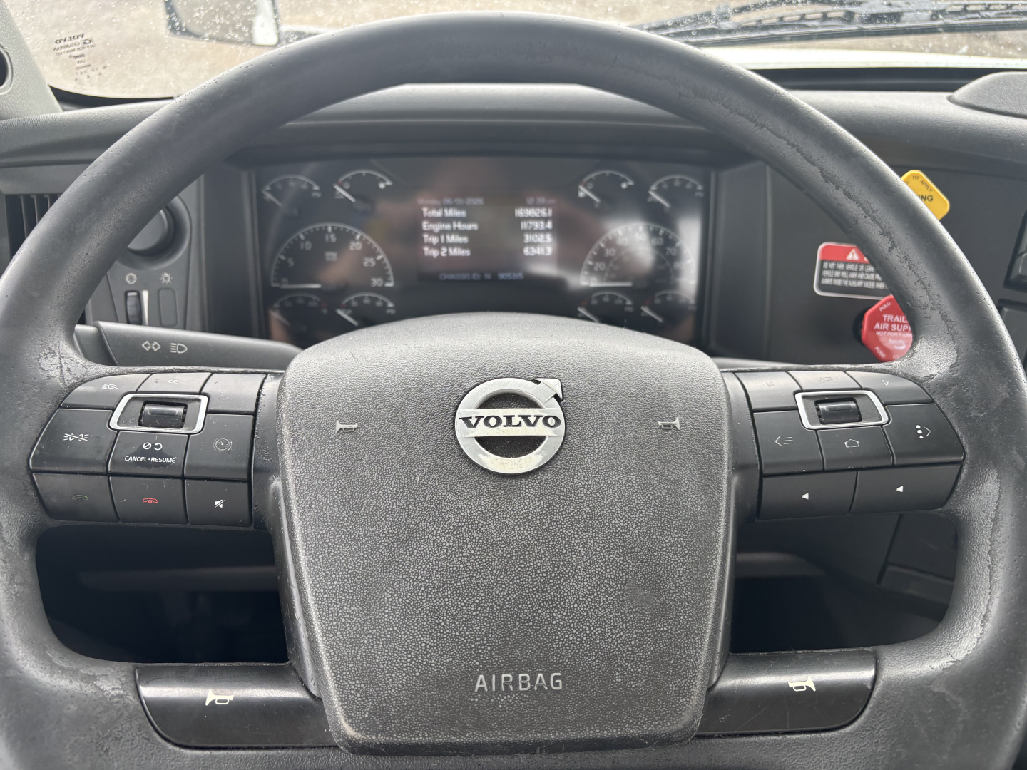 Volvo truck cockpit with steering wheel and instrument cluster. Center shows Volvo emblem on airbag cover; surrounding controls on both sides of wheel; dashboard gauges and digital display reading; warning labels on right.