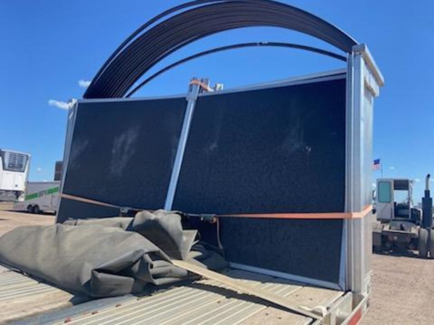 Flatbed trailer with tall metal side panels and a large coiled pipe or hose on top; rear ramp area visible; wheel wells and straps securing fabric cover in foreground.