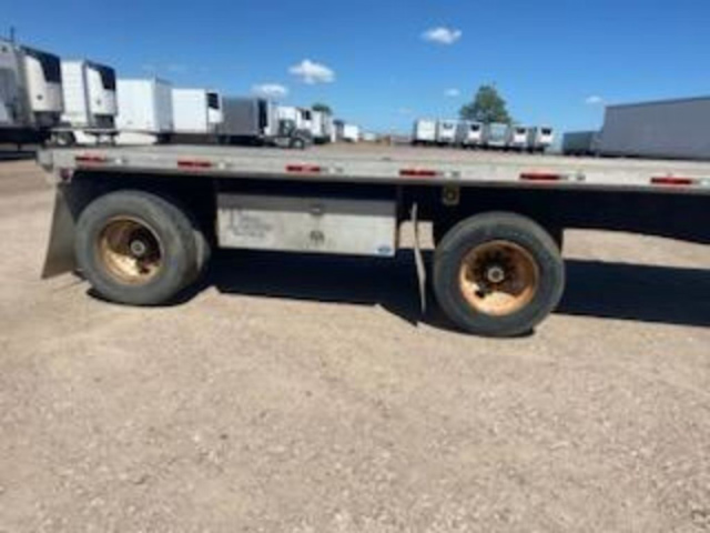 Trailer side view showing dual rear axles of a flatbed trailer with some rusted wheels and a metal toolbox; no legible text visible on components.