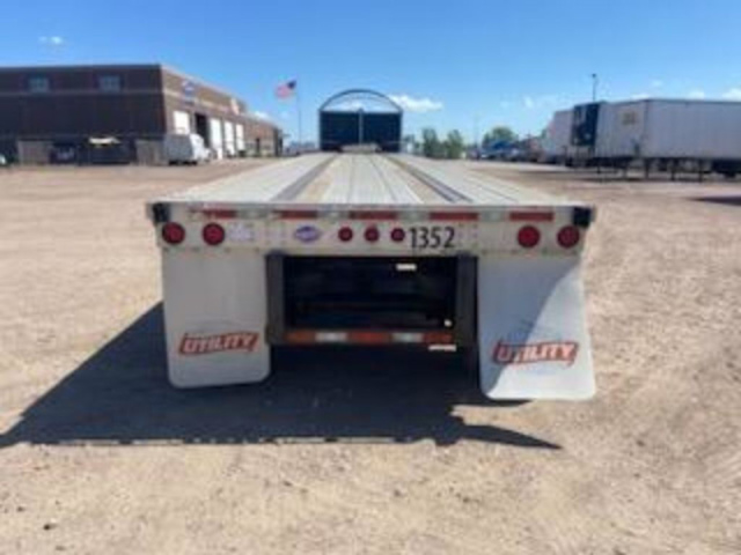 Flatbed trailer rear view showing taillights and mud flaps with 'Utility' branding and number 1352, gravel lot, other trailers in background.