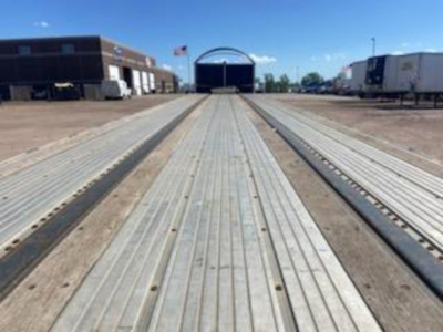 Flatbed trailer deck with long galvanized flooring and side rails, warehouse backdrop, blue sky.
