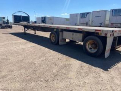 Flatbed trailer in-service showing long aluminum deck with dual rear axles and white undercarriage; no legible text visible on side; background shows equipment and clear sky.