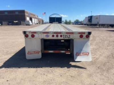 Flatbed trailer rear view showing taillights and mud flaps with 'Utility' branding and number 1352, gravel lot, other trailers in background.