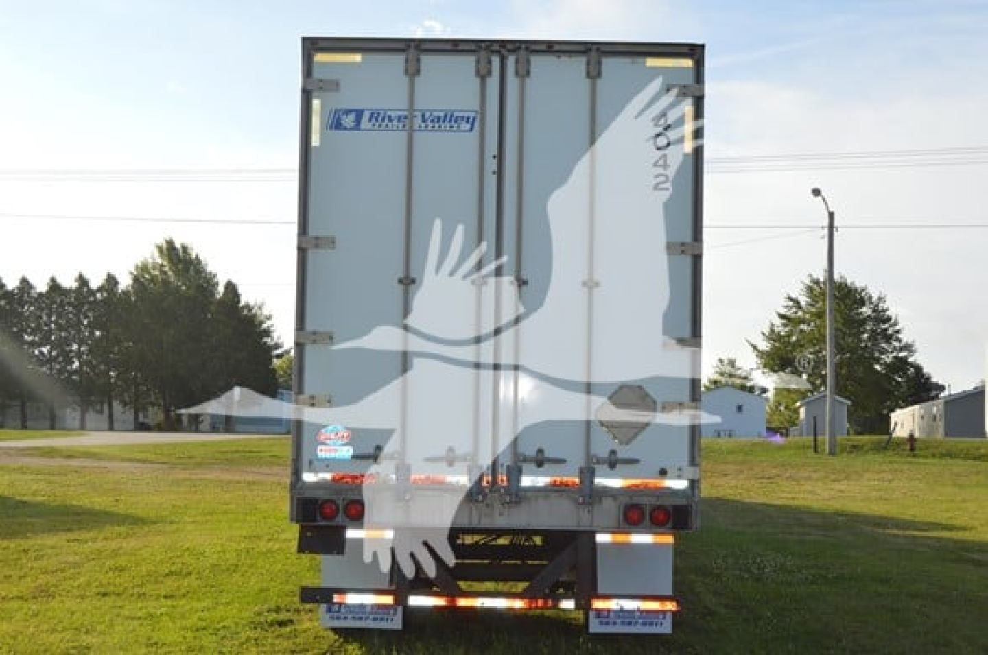 Trailer back view showing large white bird graphic on doors, River Valley logo, taillights, rear bumper, and dirt road surroundings.
