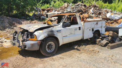 Ford F-250 Super Duty utility truck in salvage yard, front damaged with exposed engine, side utility bed, dirt ground, pile of scrap in background.