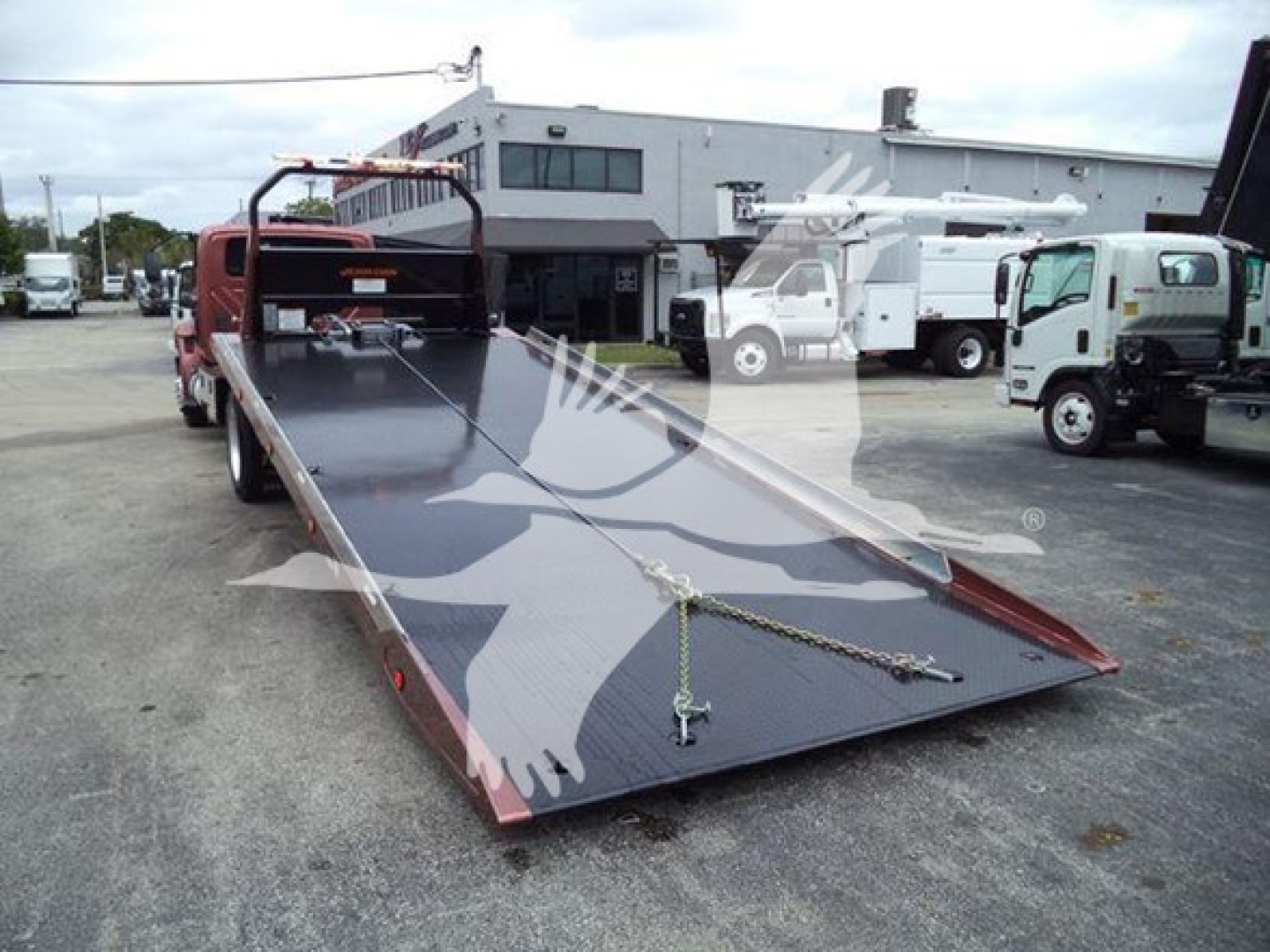Flatbed tow truck with hydraulic rollback trailer in a lot; shows metal deck with chain tie-downs and ramp; rear of cab visible
