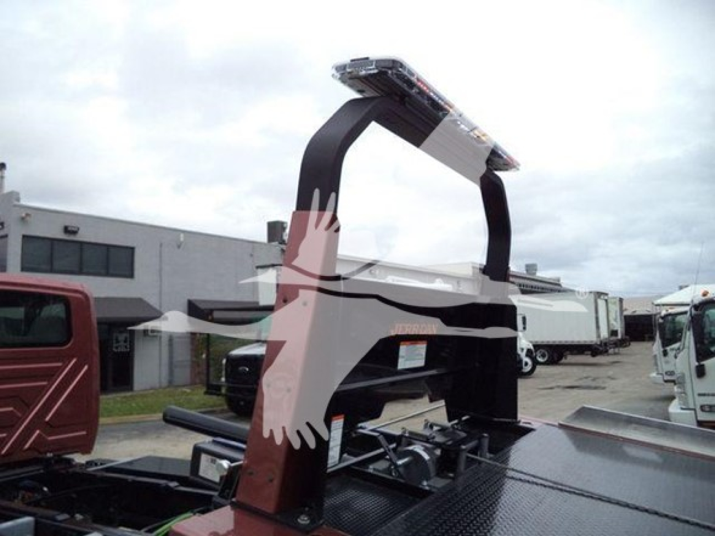 Truck bed with a heavy-duty rollback hoist arching over; no legible brand text visible; surroundings show a vehicle lot with other trucks.