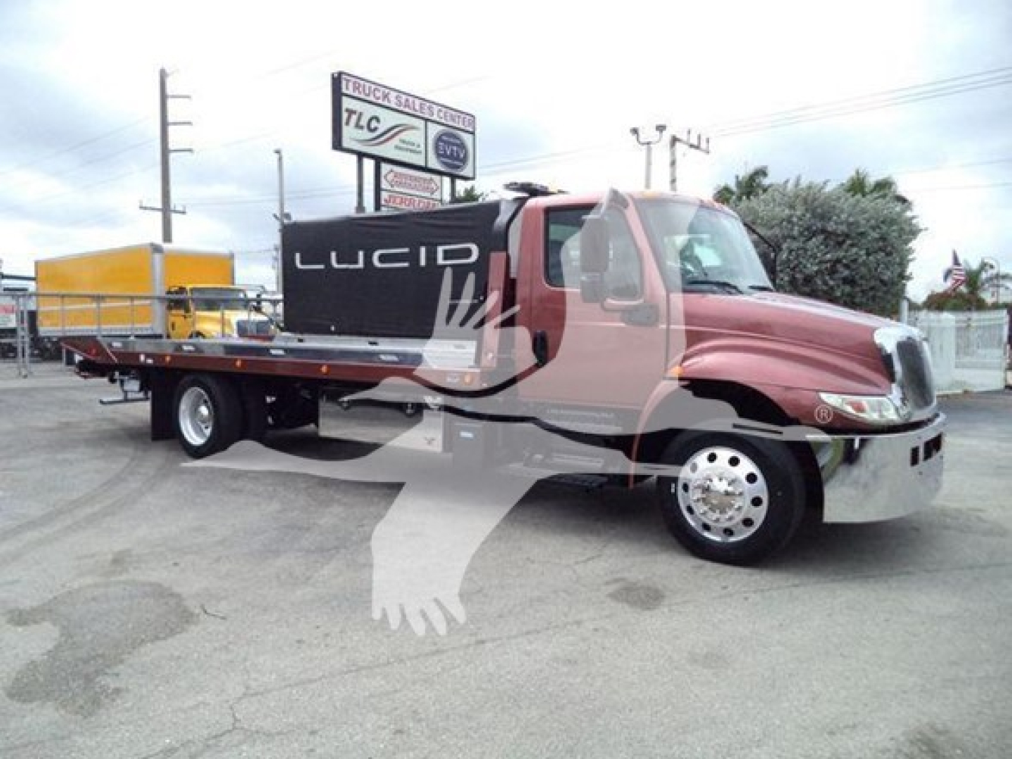 Truck with rollback bed showing LUCID on the side of the cab and a yellow container on the flatbed; dealership sign in background.