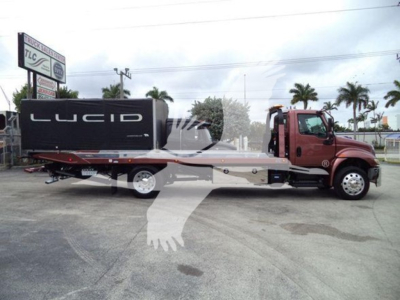 Truck with a red tow truck cab and a flatbed rollback trailer carrying a Lucid battery module on the bed, side view.