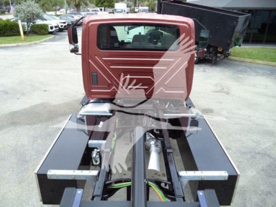 Truck cab rear view showing chassis and hydraulic components in a rollback truck. Visible components include a black frame, green and yellow wires, and a red cab with rear window.