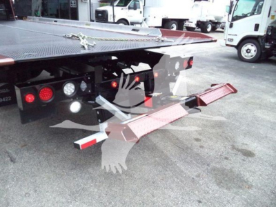 Tow truck/wrecker bed with rear ramps and tail lights visible, black metal platform, hydraulic system, and wheel chocks; International MV vehicle in background.