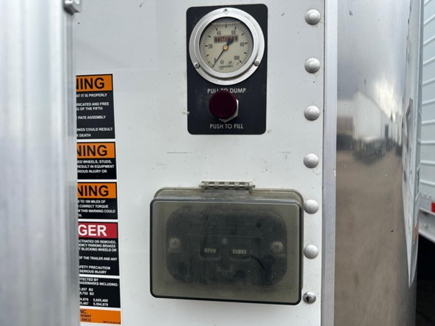 Trailer control panel of a 2024 Timpte Super Hopper showing a gauge, push to fill button, and a metal access panel with labels and rivets.