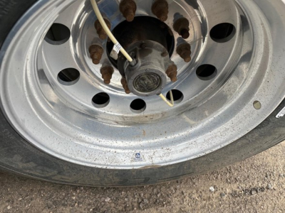 Tire and wheel of a 2024 Timpte Super Hopper trailer hub area with lug nuts and hub cover; visible alloy wheel with rust spots on lug nuts and a white cable tied around near center.