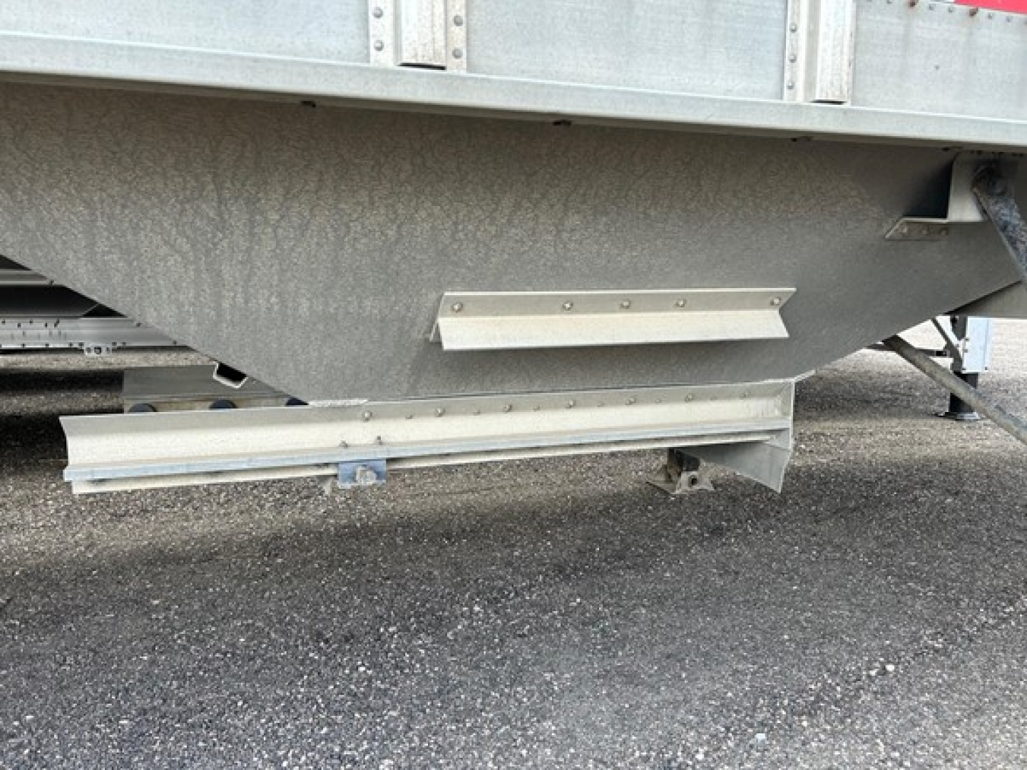 Trailer underside showing grain hopper with metal support brackets and reflective strip, gravel ground.