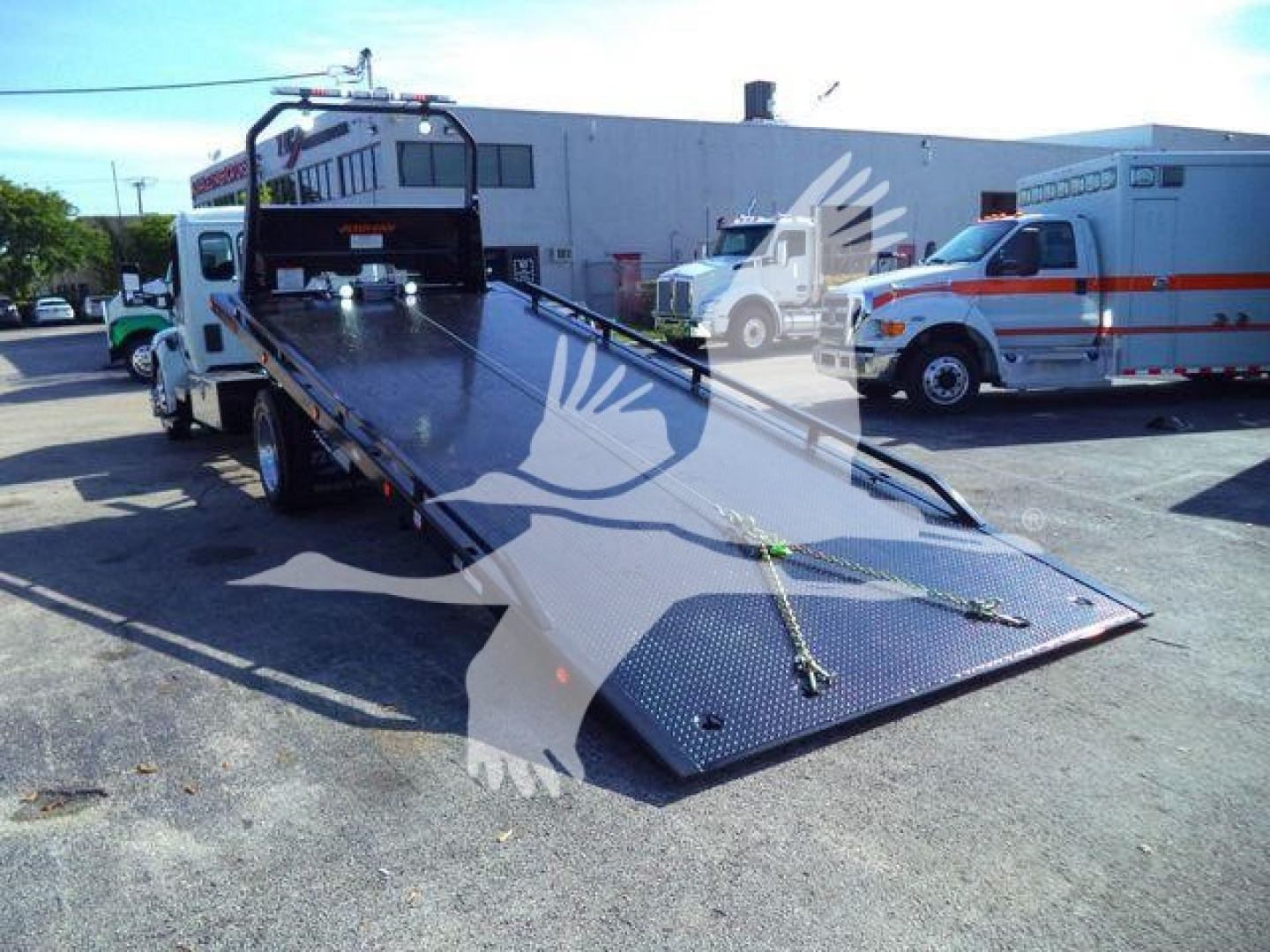 Peterbilt 536 rollback tow truck with extended flatbed in view, white cab, black bed, emergency vehicle in background, stock number 1PETE536XLP22.