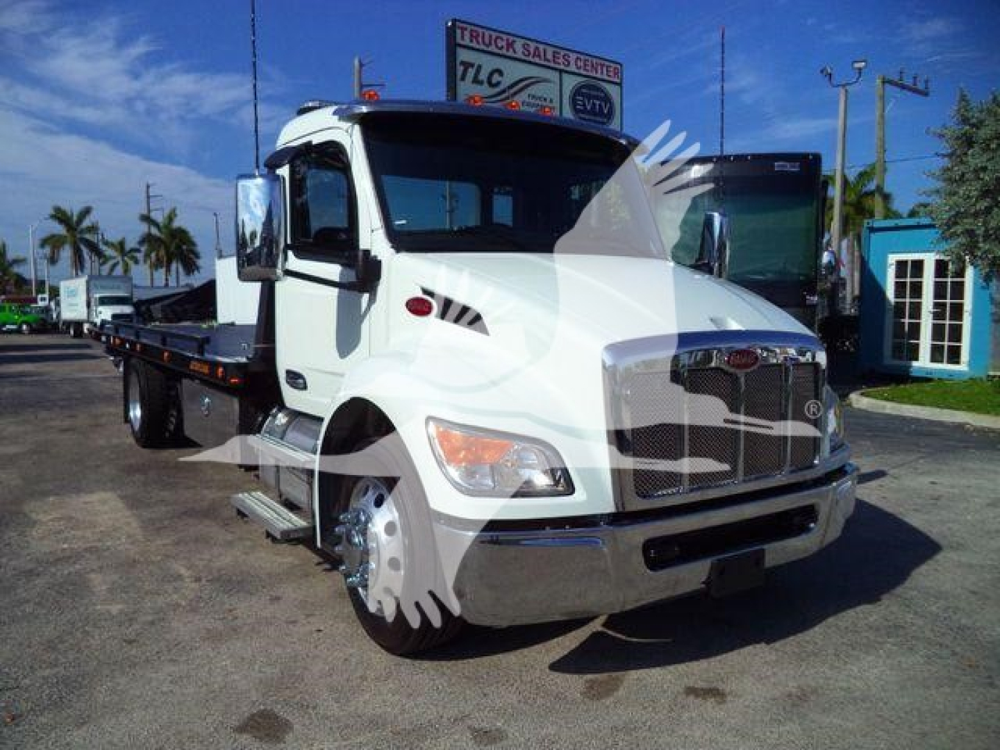 Truck: 2026 Peterbilt 536 rollback truck with a flatbed, white cab, chrome grille, dual side mirrors, and stacked wheels. Visible text on signs in background is blurry.