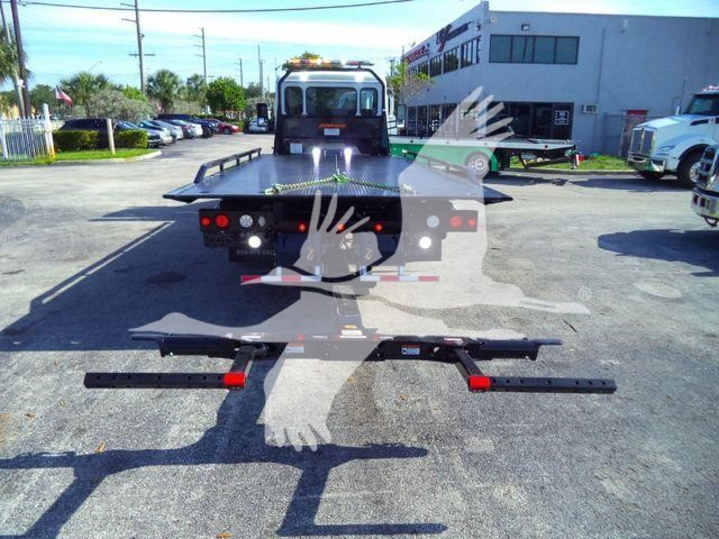 Back of a Peterbilt rollback truck with metal steps and rear bumper pieces laid on pavement; dealership lot in background.