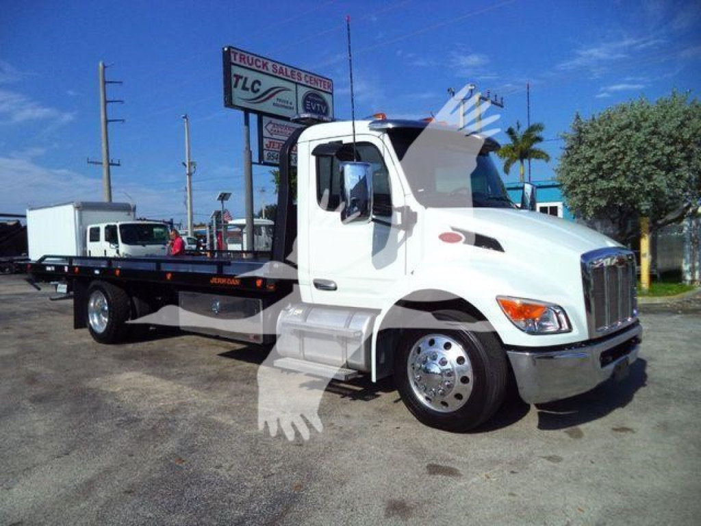 Peterbilt 536 rollback tow truck in white with black flatbed, front cab with chrome grille, sign in background. Visible text on sign includes truck sales center TLC and others.