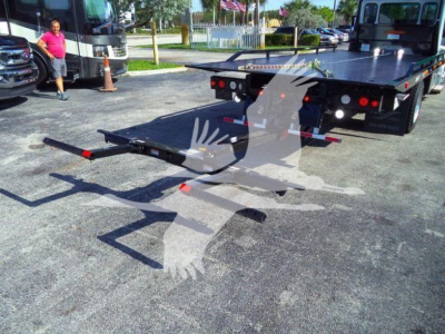 Flatbed rollback truck with loading ramp extended showing rear view and wheels, Peterbilt signage and dealership lot in background.