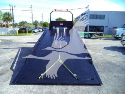 Truck bed of a 2026 Peterbilt 536 rollback showing a long metal loading ramp with a front winch and safety chains.