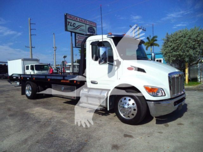 Peterbilt 536 rollback tow truck in white with black flatbed, front cab with chrome grille, sign in background. Visible text on sign includes truck sales center TLC and others.