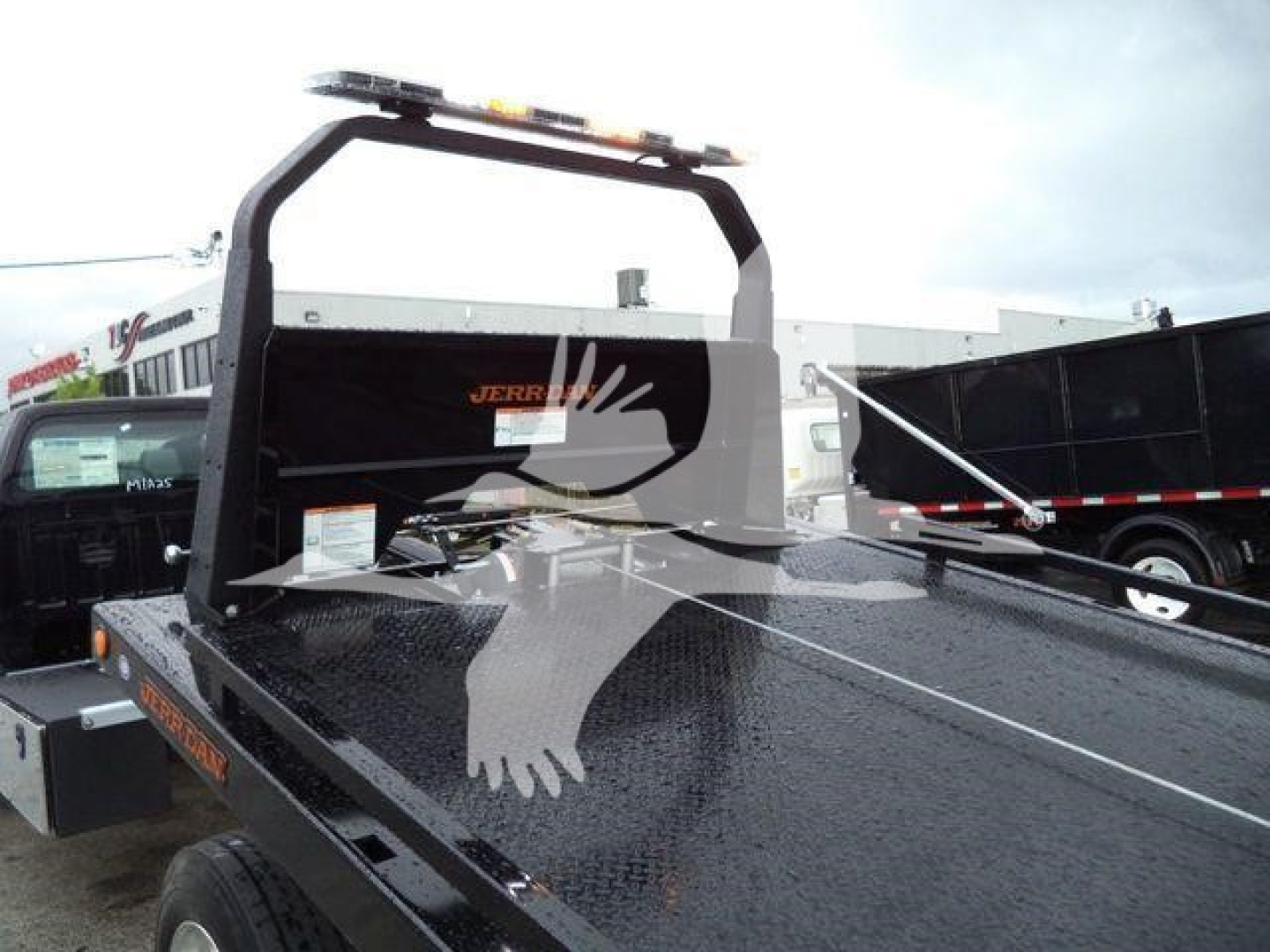 Flatbed tow truck bed with black diamond plate and rollover pickup rack. Visible branding JBERT? and orange warning stickers; light bar on cab frame.