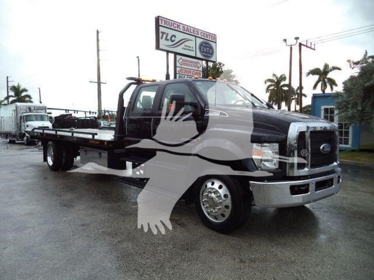 Ford F-750 rollback tow truck with flatbed in a dealership lot. Visible text on signboard: TRUCK SALES CENTER, 7LC; license plate not legible. The truck features a large front chrome bumper, dual rear wheels, and a black crew cab.