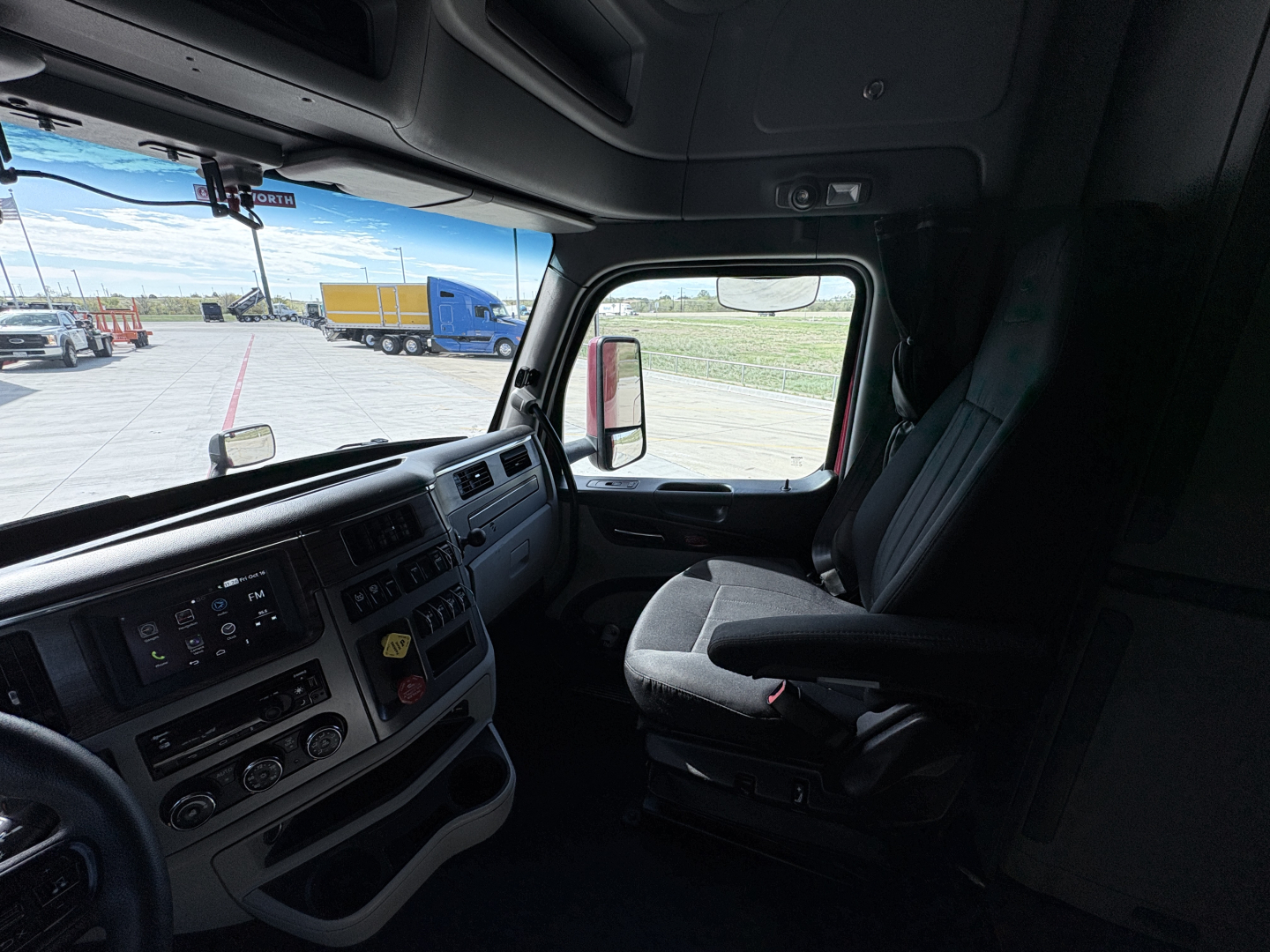 Truck cab interior of a 2023 Peterbilt 579 showing dashboard controls, steering wheel, and driver's seat. Exterior view through window with other trucks in lot.