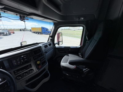 Truck cab interior of a 2023 Peterbilt 579 showing dashboard controls, steering wheel, and driver's seat. Exterior view through window with other trucks in lot.