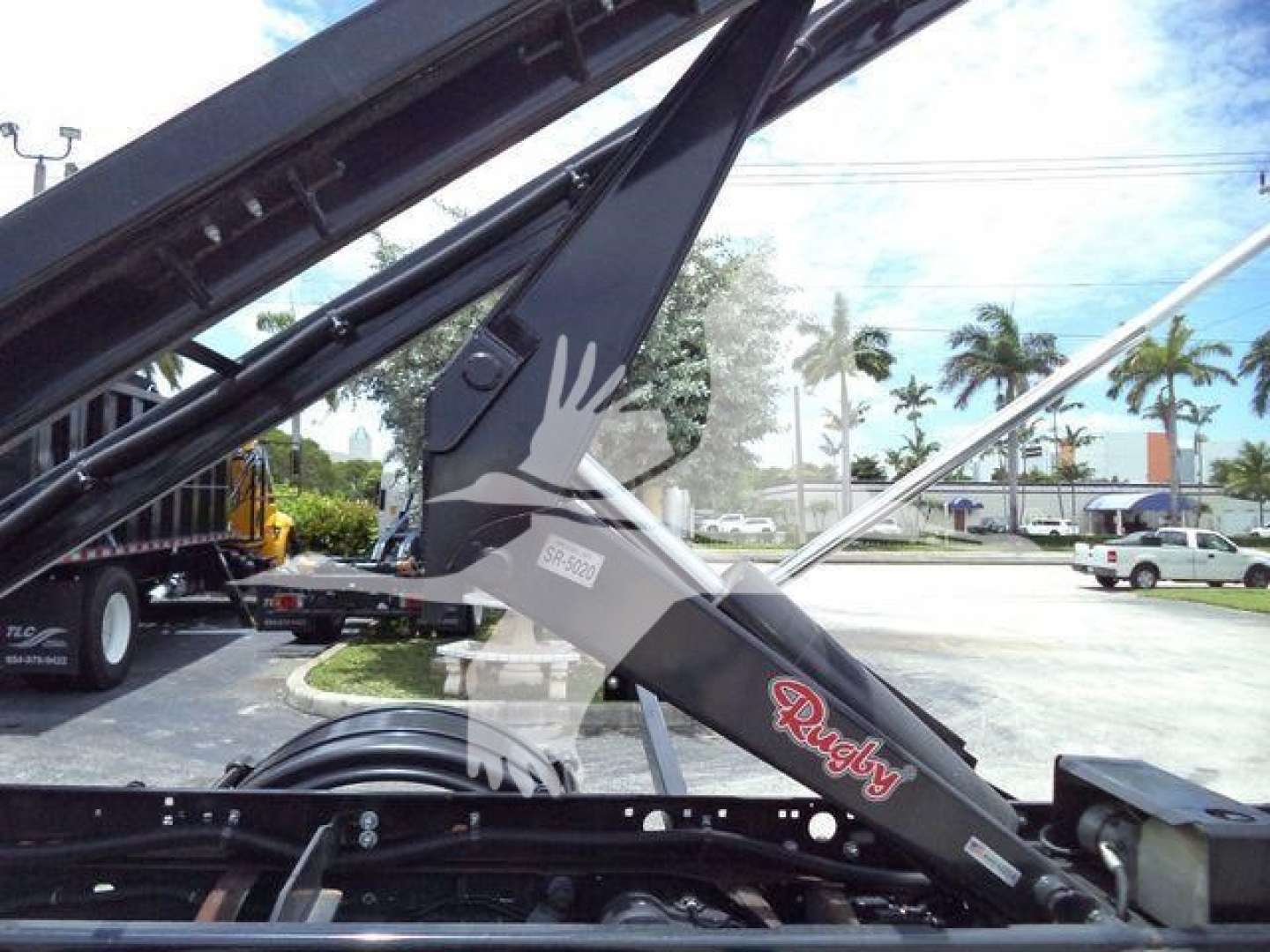 Ram 5500 roll-off truck component with hydraulic arm and Rugby branding visible; labels on the arm show model/serial text.