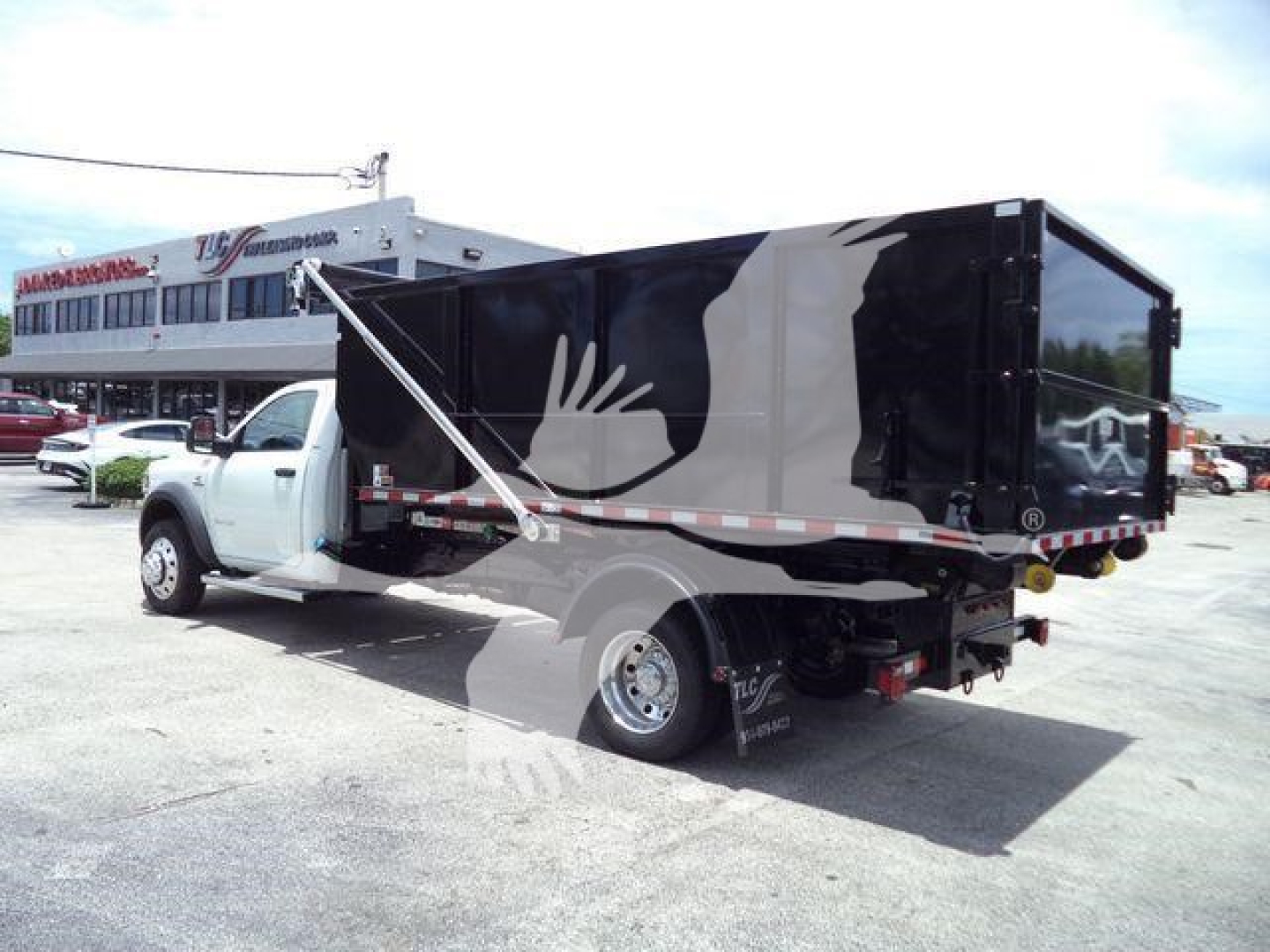 Ram 5500 truck with a roll-off bed and black/gray graphic, side-dump configuration visible, rear view.
