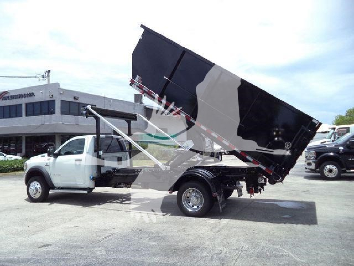 Ram 5500 SLT roll-off truck with hydraulic dump bed in raised position, side view showing cab, white body, black dump bed, dealership lot in background.