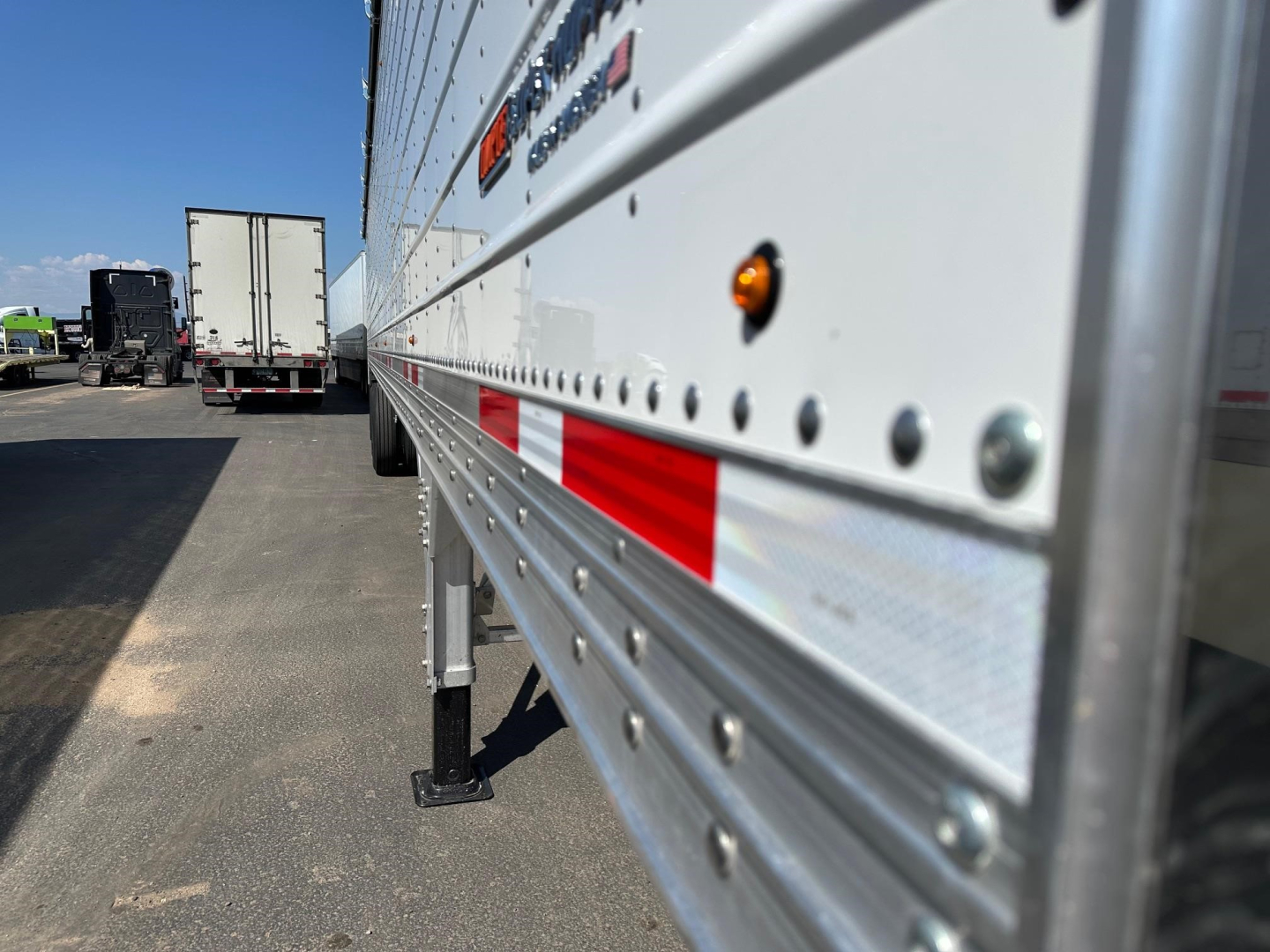 Trailer side of a 2026 Timpte Super Hopper showing riveted aluminum side with red reflective stripes and orange marker lights; parking lot background with other trailers.