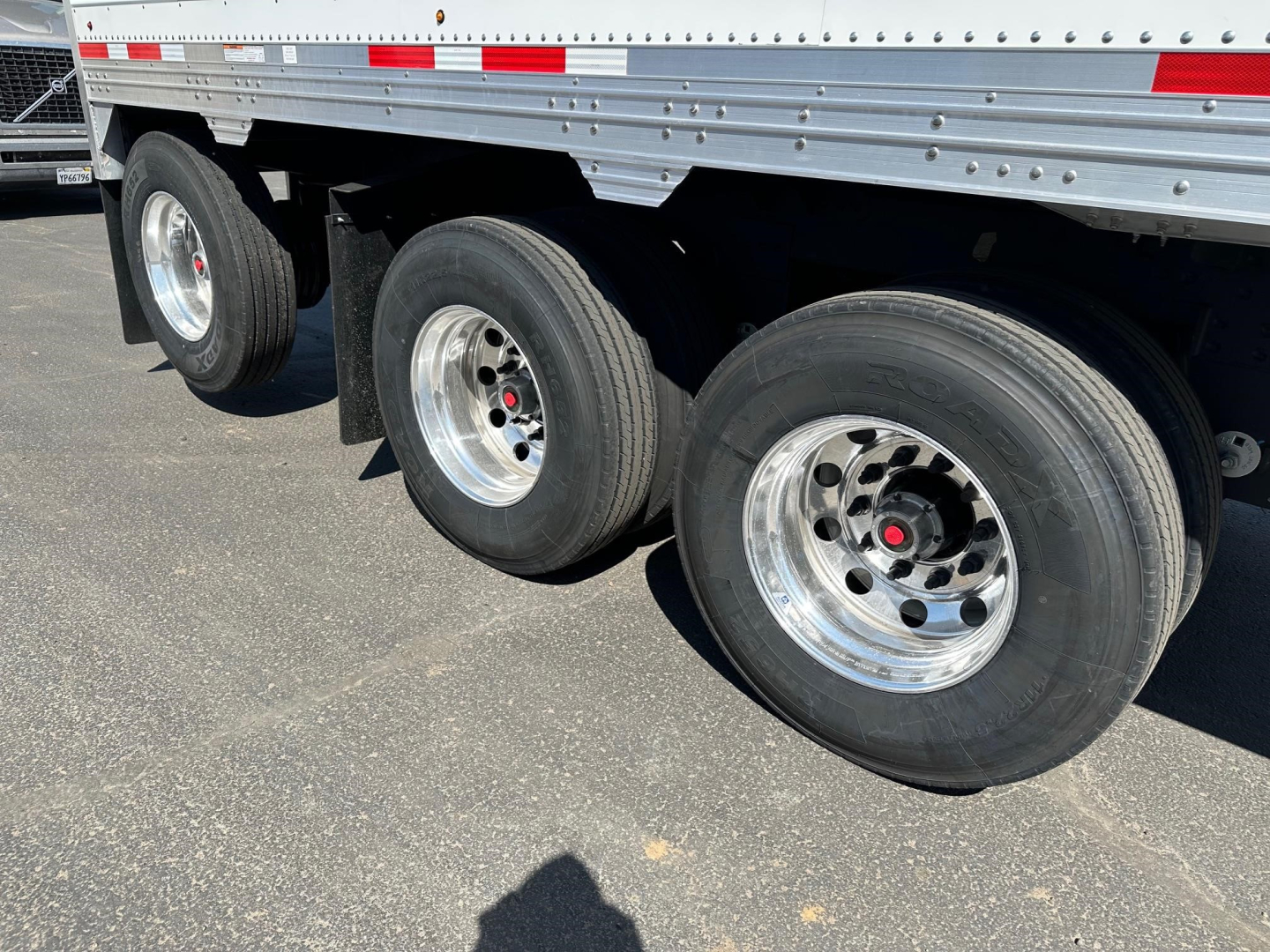 Three axles of a hopper trailer with polished aluminum rims and large tires, side rails and reflective tape along top edge.