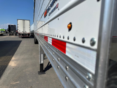 Trailer side of a 2026 Timpte Super Hopper showing riveted aluminum side with red reflective stripes and orange marker lights; parking lot background with other trailers.