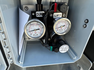 Two pressure gauges labeled DUMP and LIFT in a Timpte Super Hopper trailer control panel with manuals and cables visible.