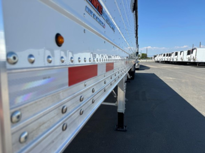 Semi trailer side panel with riveted aluminum siding, orange marker light, red and white reflective tape, and support leg visible near the wheel. Row of similar trailers in the background.