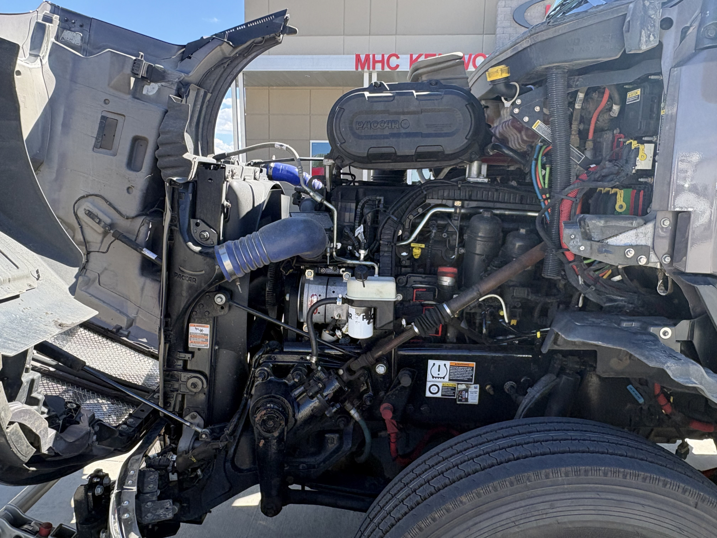 Kenworth T680 engine bay showing diesel engine with hoses, wiring, and open hood; visible air intake hose, coolant reservoir cap, and various labels.