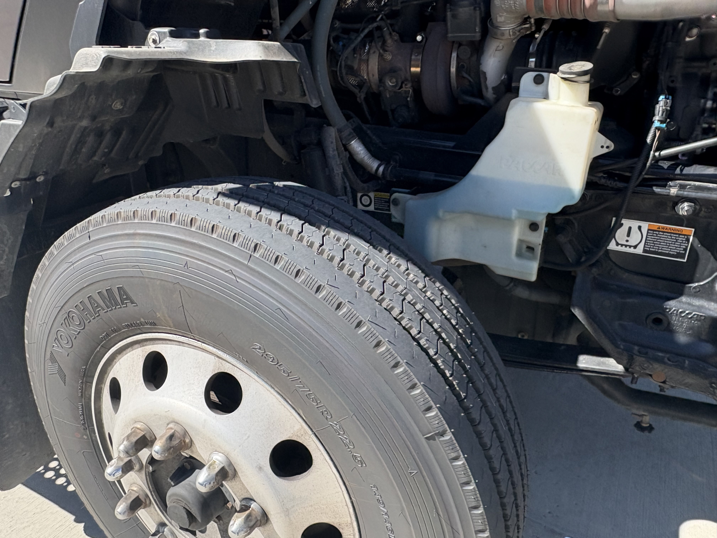 Kenworth T680 engine bay side showing front tire with Yokohama tire and wheel, coolant reservoir, battery area, and engine components visible.