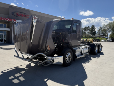 Kenworth T680 conventional daycab tractor in a dealership lot, front section damaged with missing bumper and exposed frame; windshield intact; tandem rear axles visible.