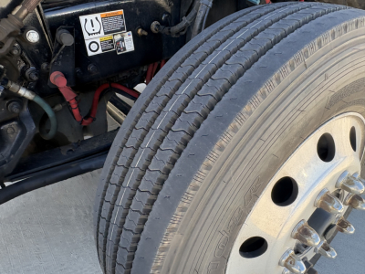 Kenworth T680 truck front wheel and engine bay view showing tire, wheel rim, and battery area with warning label.