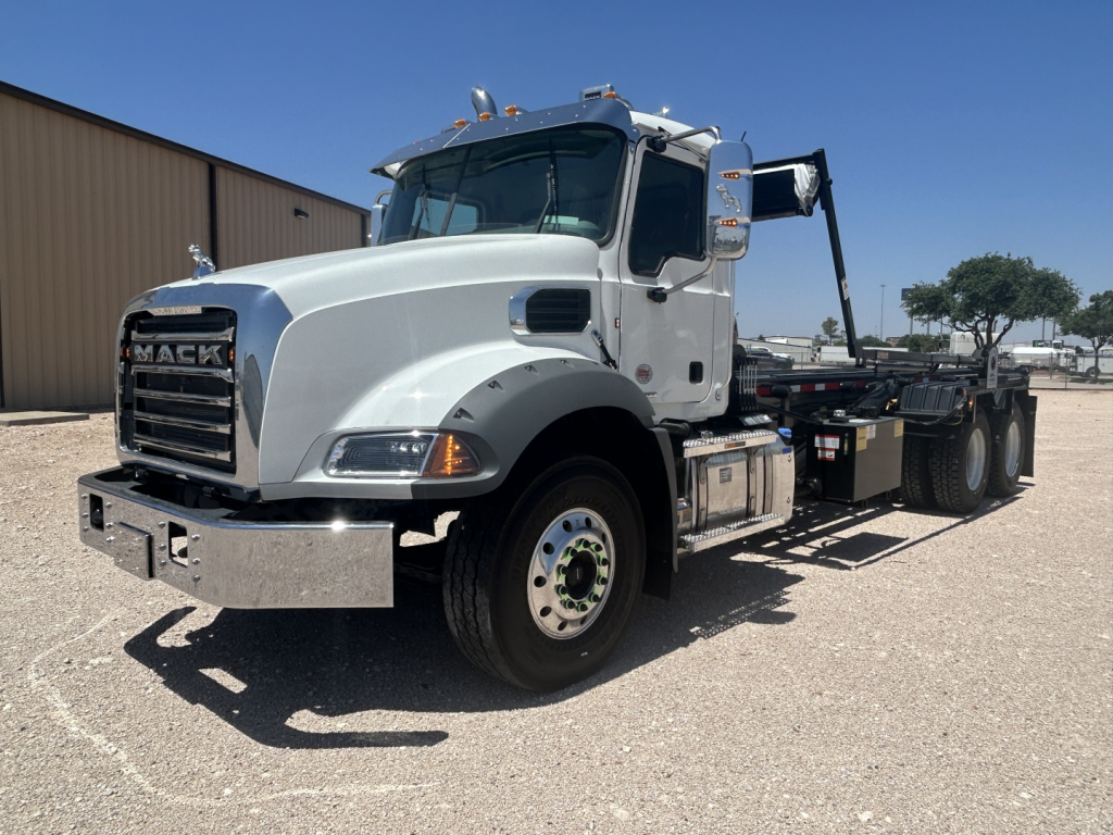 Mack GR64B truck in a dealership lot from a front-right angle. Cab and front fender visible with chrome bumper, black grille, large side mirrors, and a roll-off bed behind the cab.