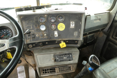 Dashboard and steering wheel area of a Mack RD688S truck cab showing gauges, switches, yellow tag, and gear shifter.