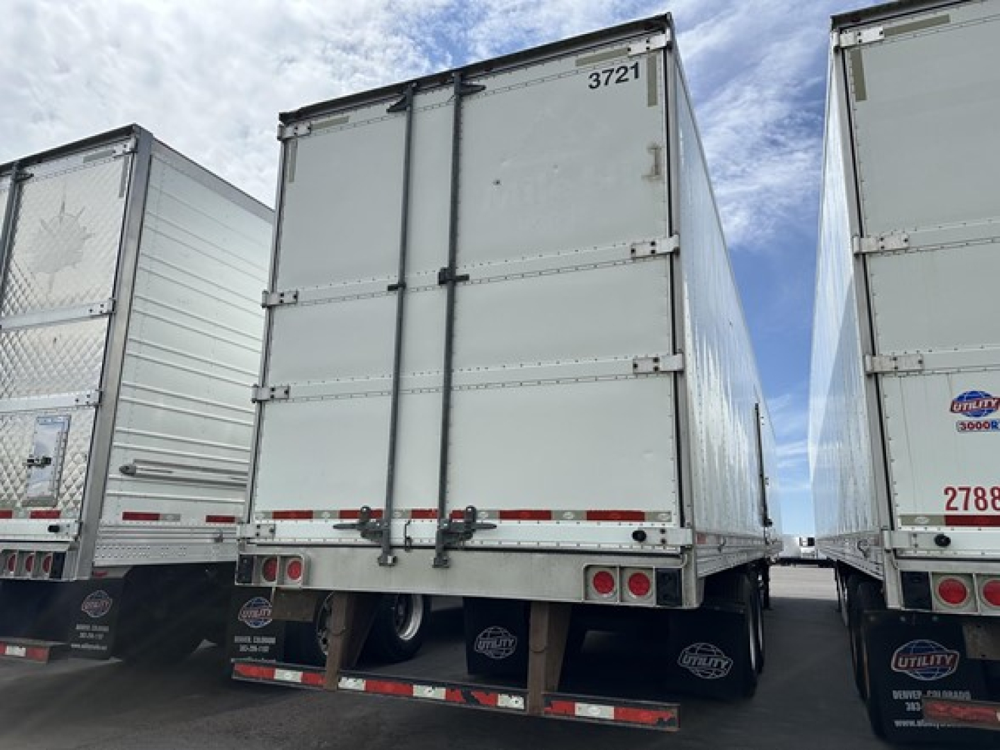 Utility trailer rear view showing hinged double doors with black latches and reflective tapes, second trailer adjacent, gray metal panels.