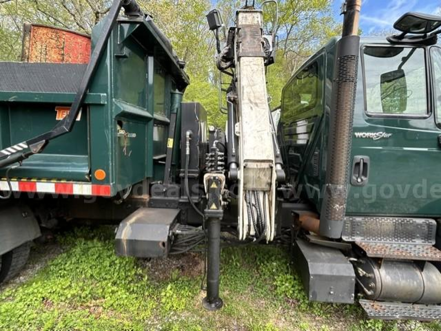 Front view of a 2012 International 7400 WorkStar dump truck with a central hydraulic crane/piler between the cab and dump bed. Visible hoses, cylinders, and stabilizer legs.