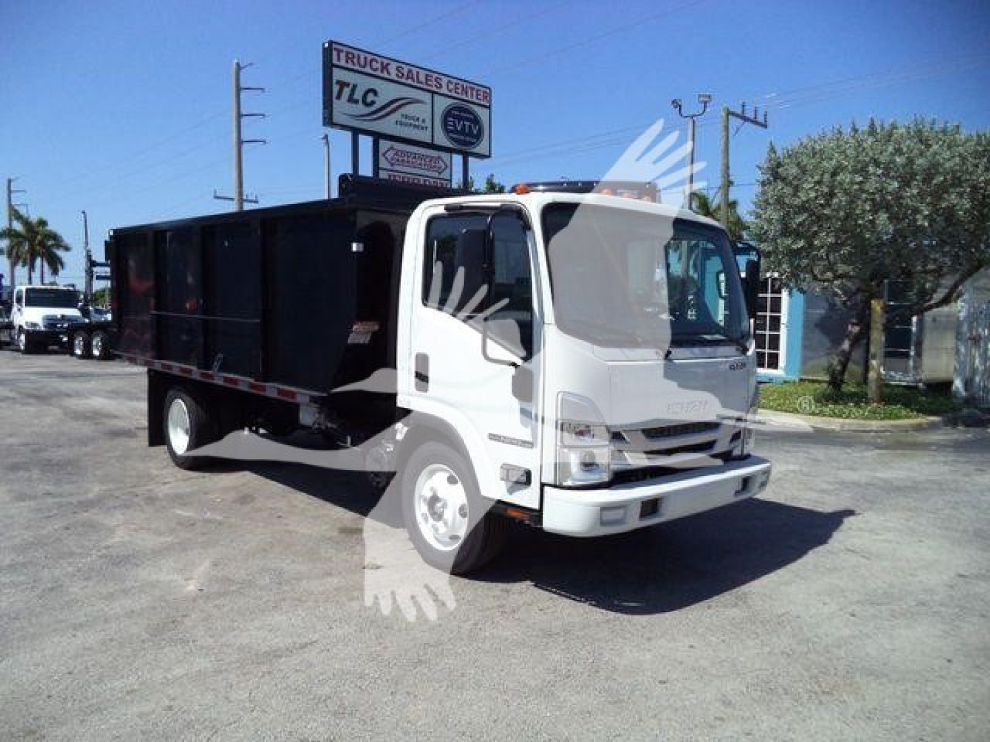 Isuzu NPR HD medium-duty truck in a lot, white cab and black dump bed; TLC Truck Sales Center sign visible in background