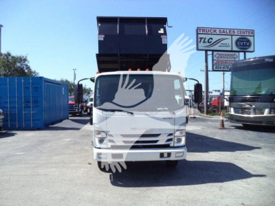 Isuzu NPR HD light truck in a lot with a raised dump bed, front view. Visible signage in background includes 'TRUCK SALES CENTER' and other signs. White cab with black dump bed raised high.